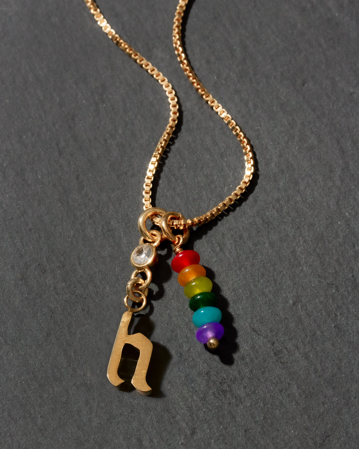 Gold necklace with rainbow beads and letter 'h' charm on a dark background