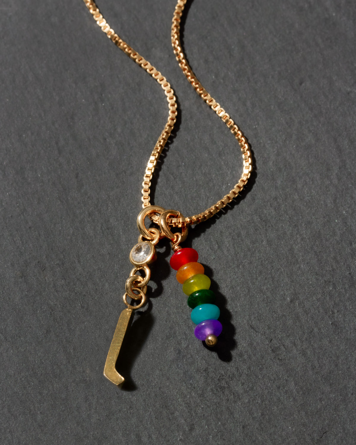 Gold necklace with rainbow charm and letter 'l' on a dark gray background