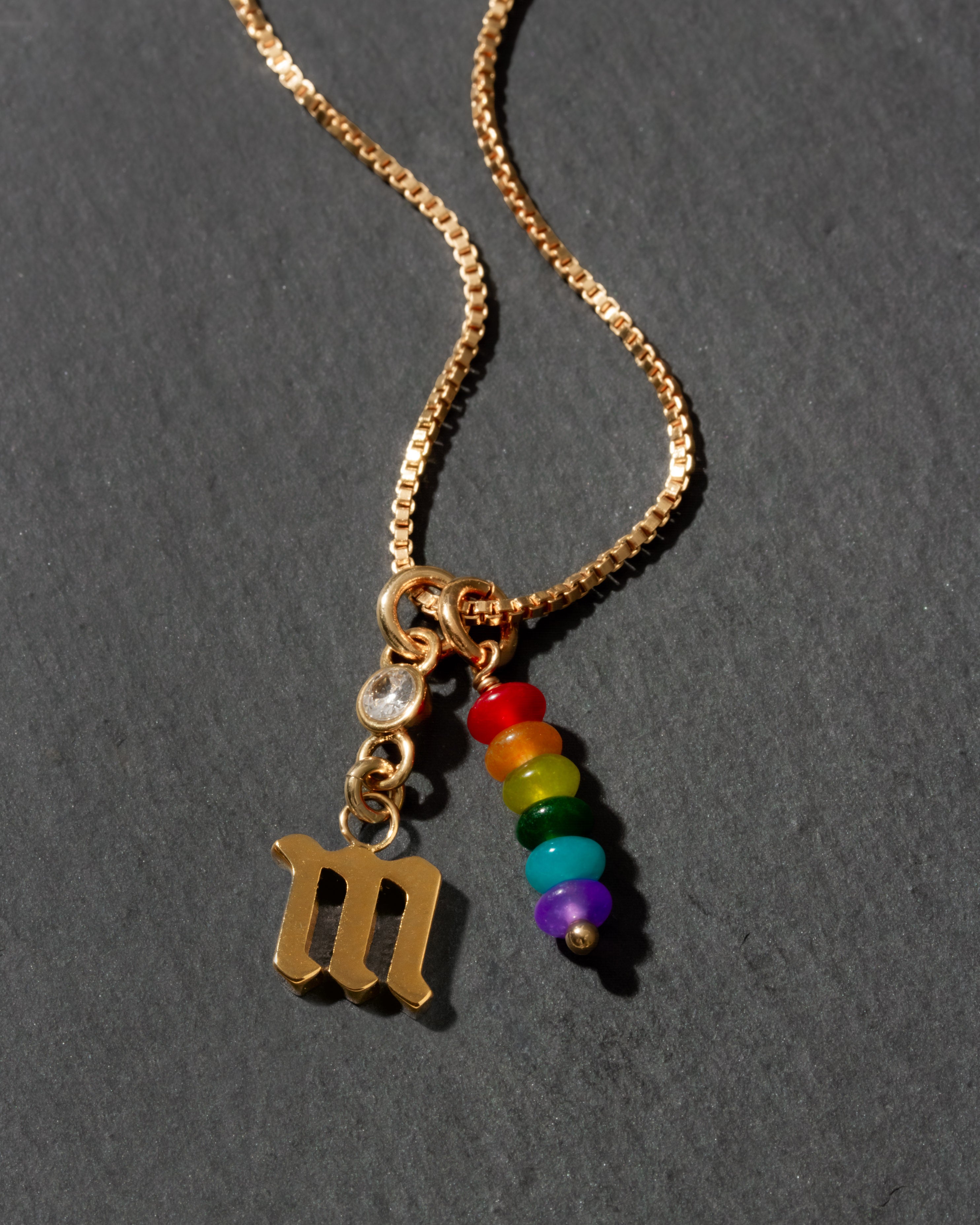 Gold necklace with a letter 'm' pendant and rainbow beads on a dark background