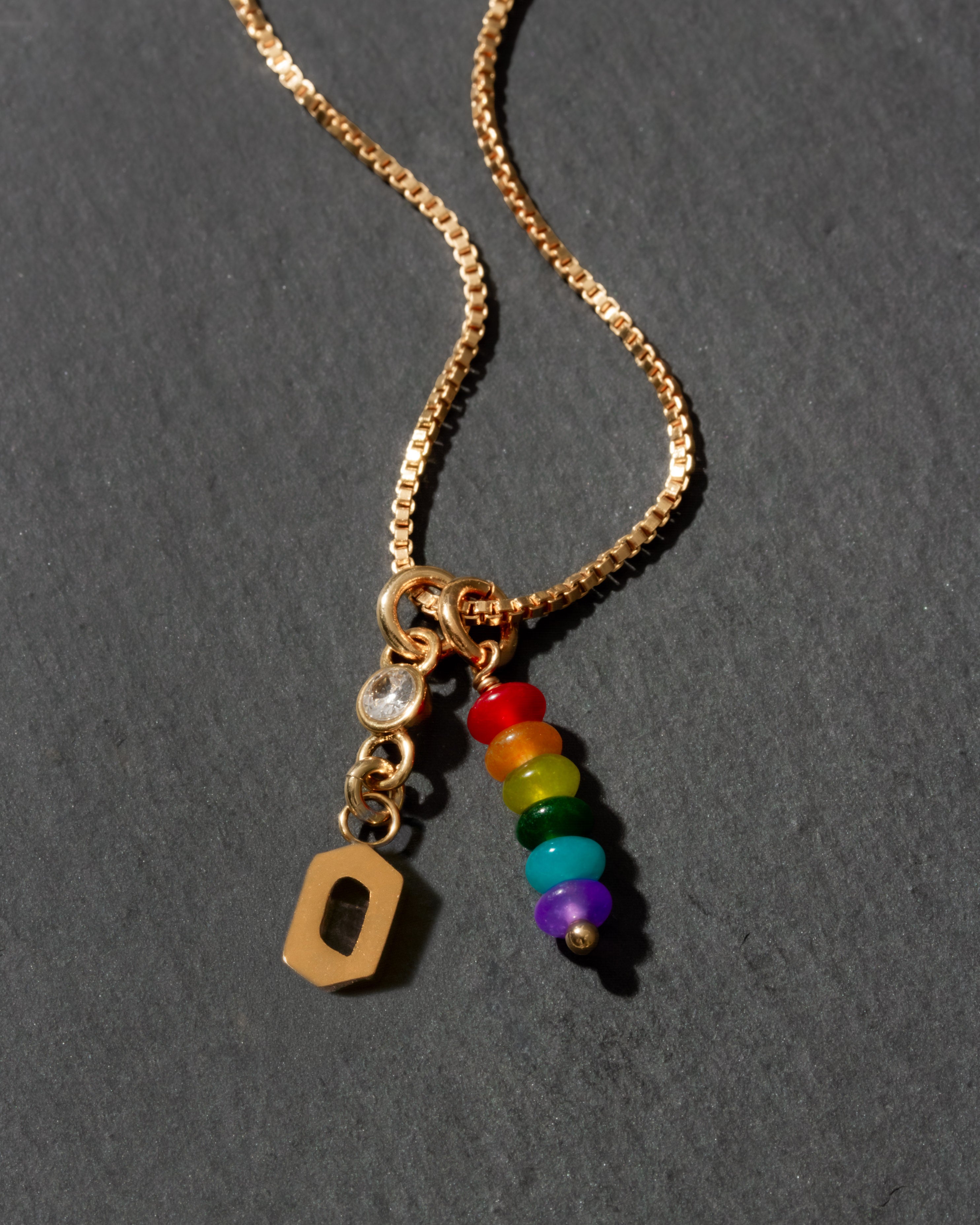 Gold necklace with rainbow charm and letter 'O' pendant on a dark background