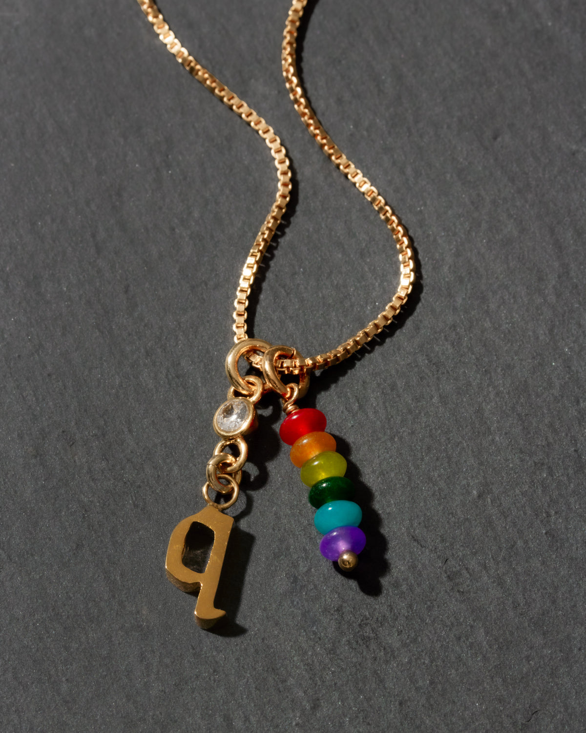 Gold necklace with rainbow charm and letter 'Q' on a dark gray background