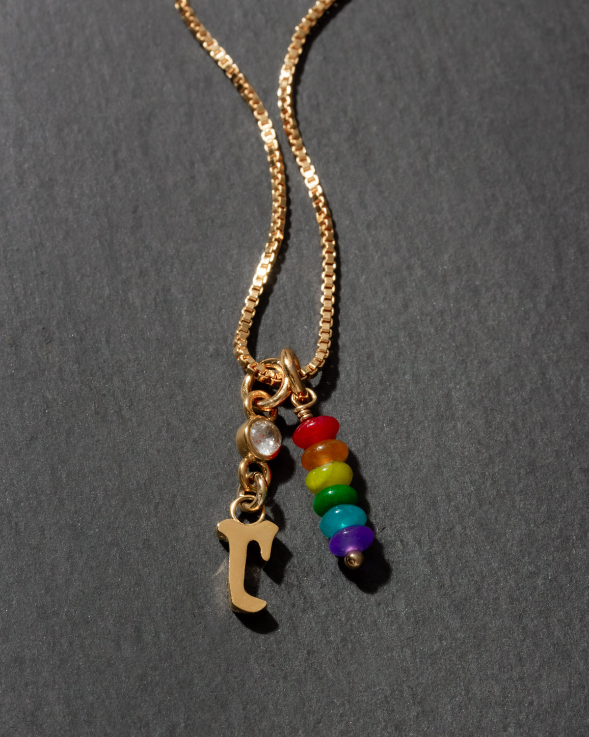 Necklace with gold chain, letter 'r' charm, and rainbow bead charms on a dark background