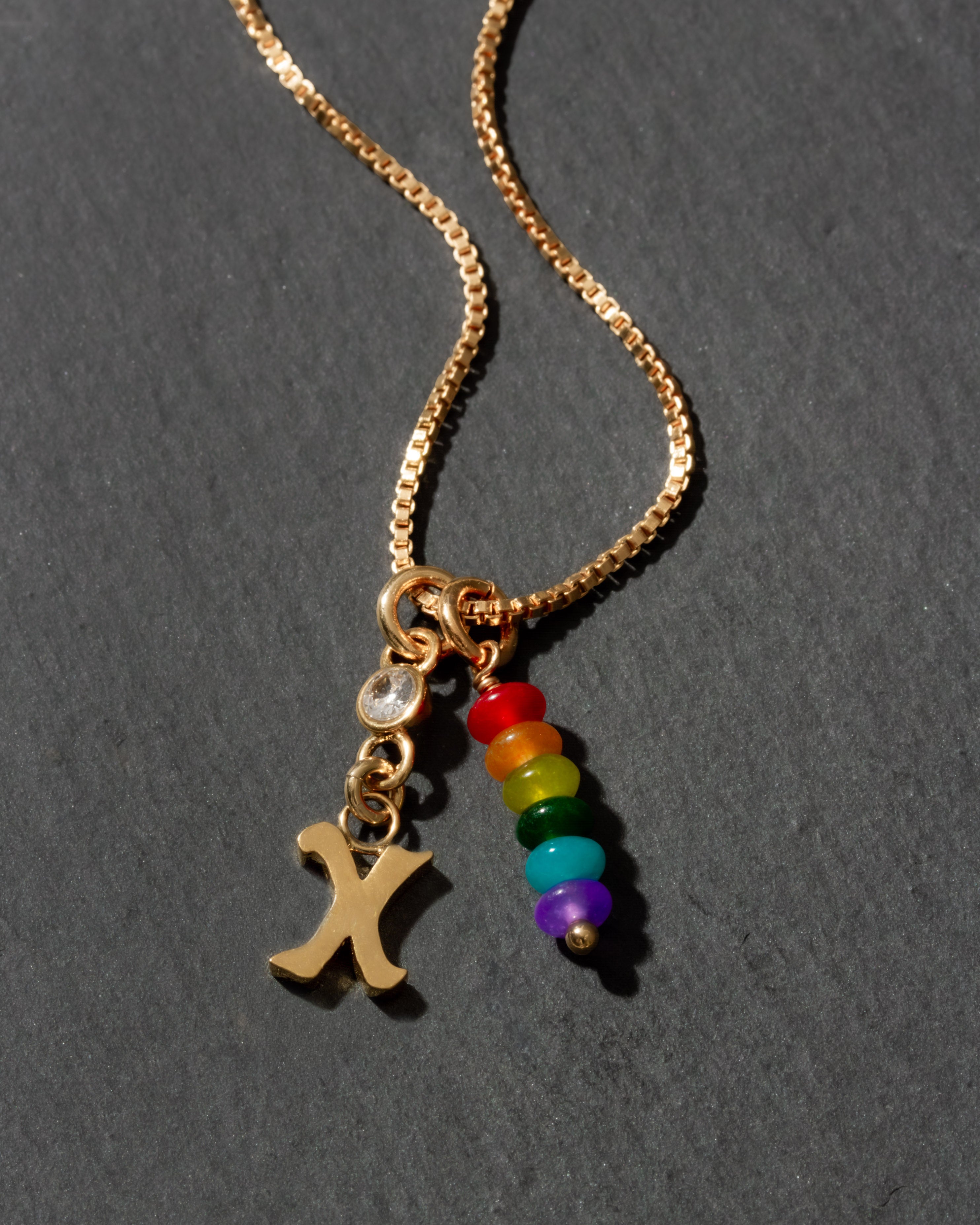 Gold necklace with rainbow charm and letter 'X' on a dark background