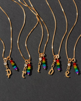 Gold initial necklaces spelling out PRIDE with rainbow charm pendants on a dark background