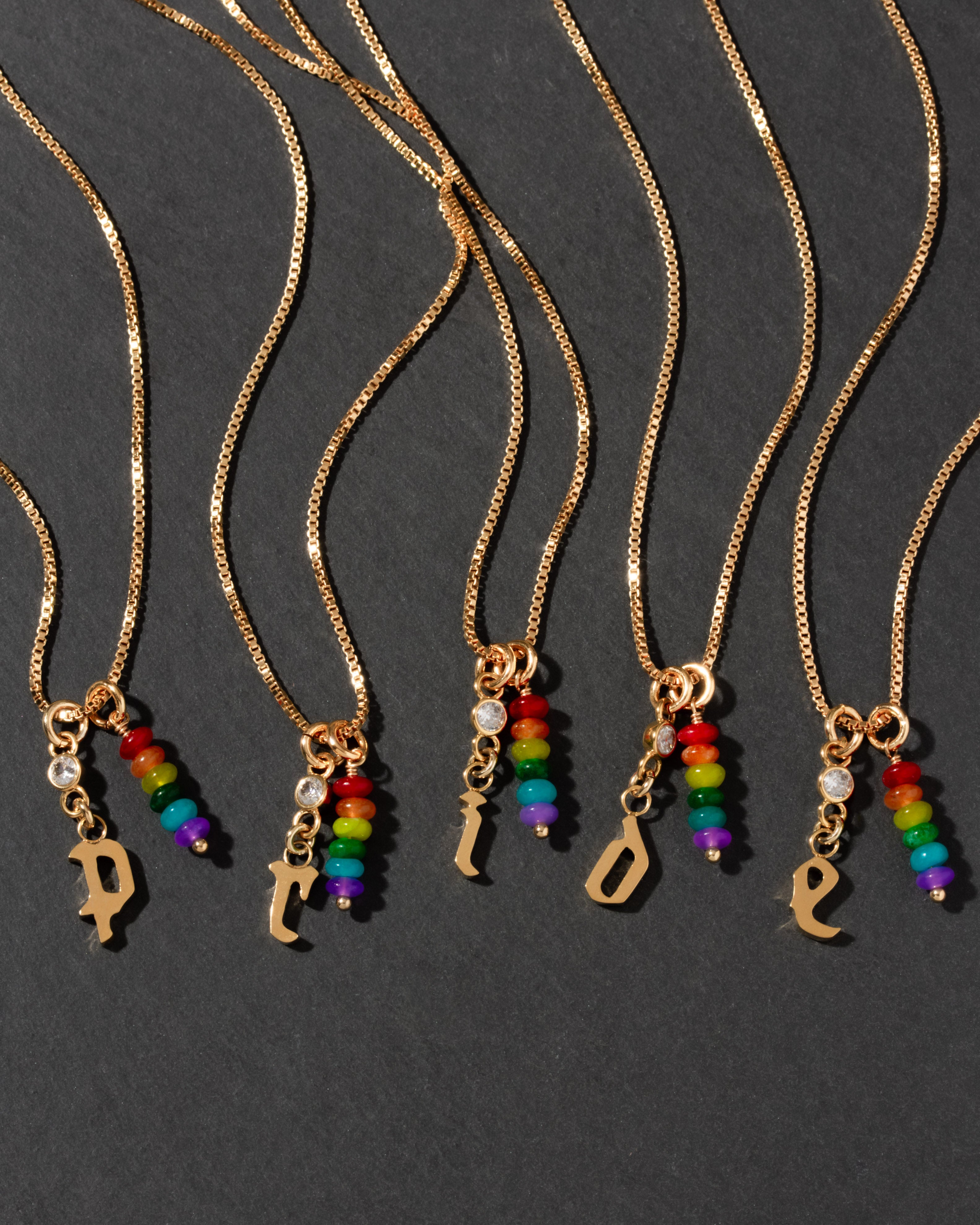 Gold initial necklaces spelling out PRIDE with rainbow charm pendants on a dark background