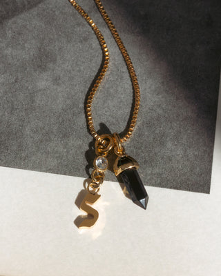 Gold necklace with a letter 's' and black crystal pendant on a gray and white background