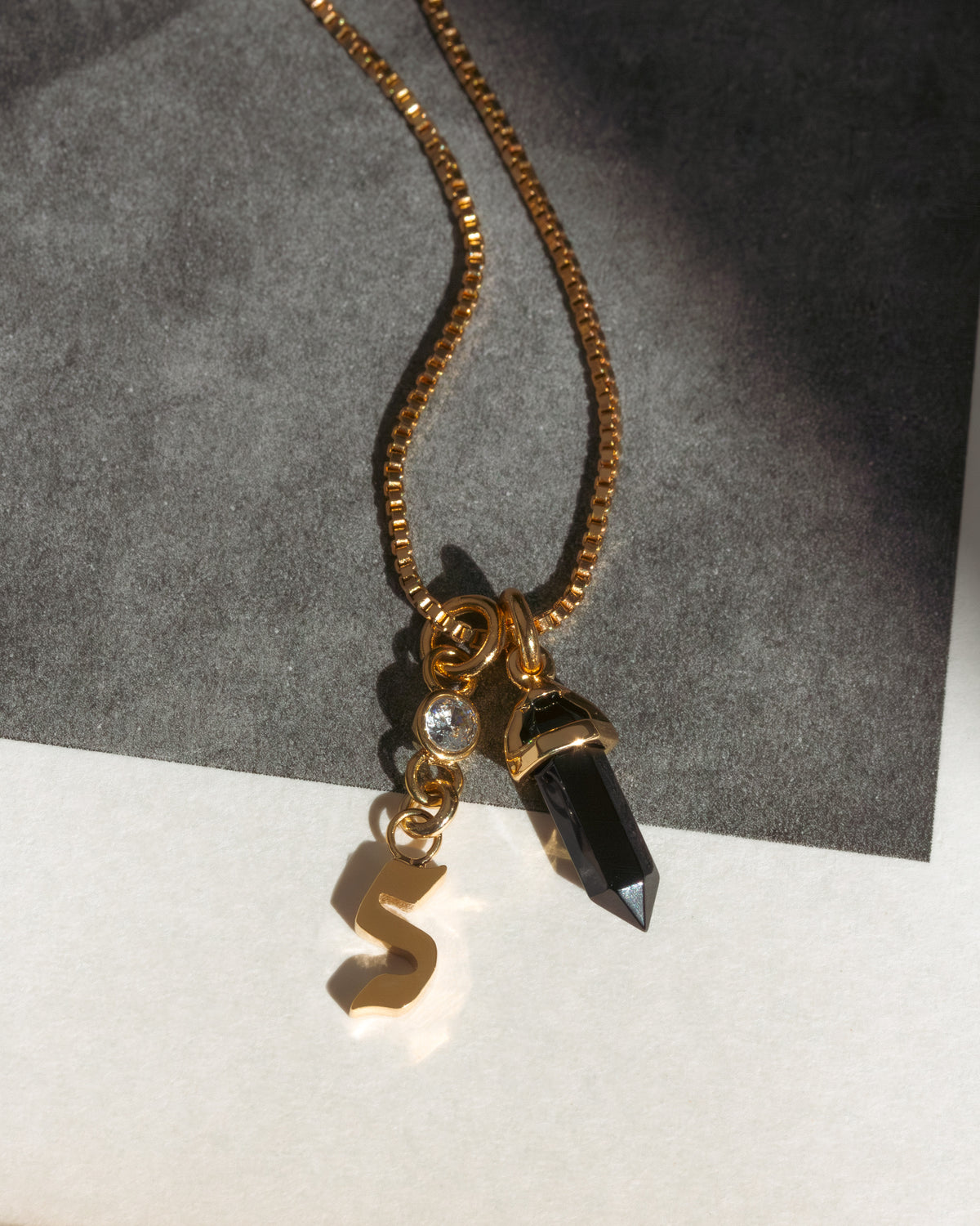 Gold necklace with a letter 's' and black crystal pendant on a gray and white background