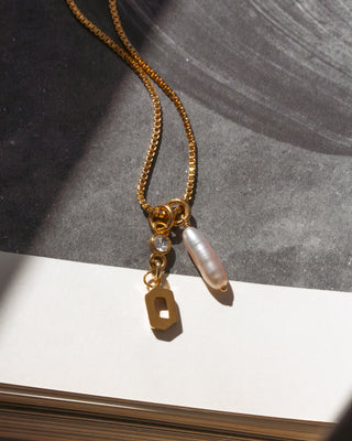 Gold necklace featuring a letter O initial charm, a sparkling CZ accent, and a luminous freshwater stick pearl on a box chain.