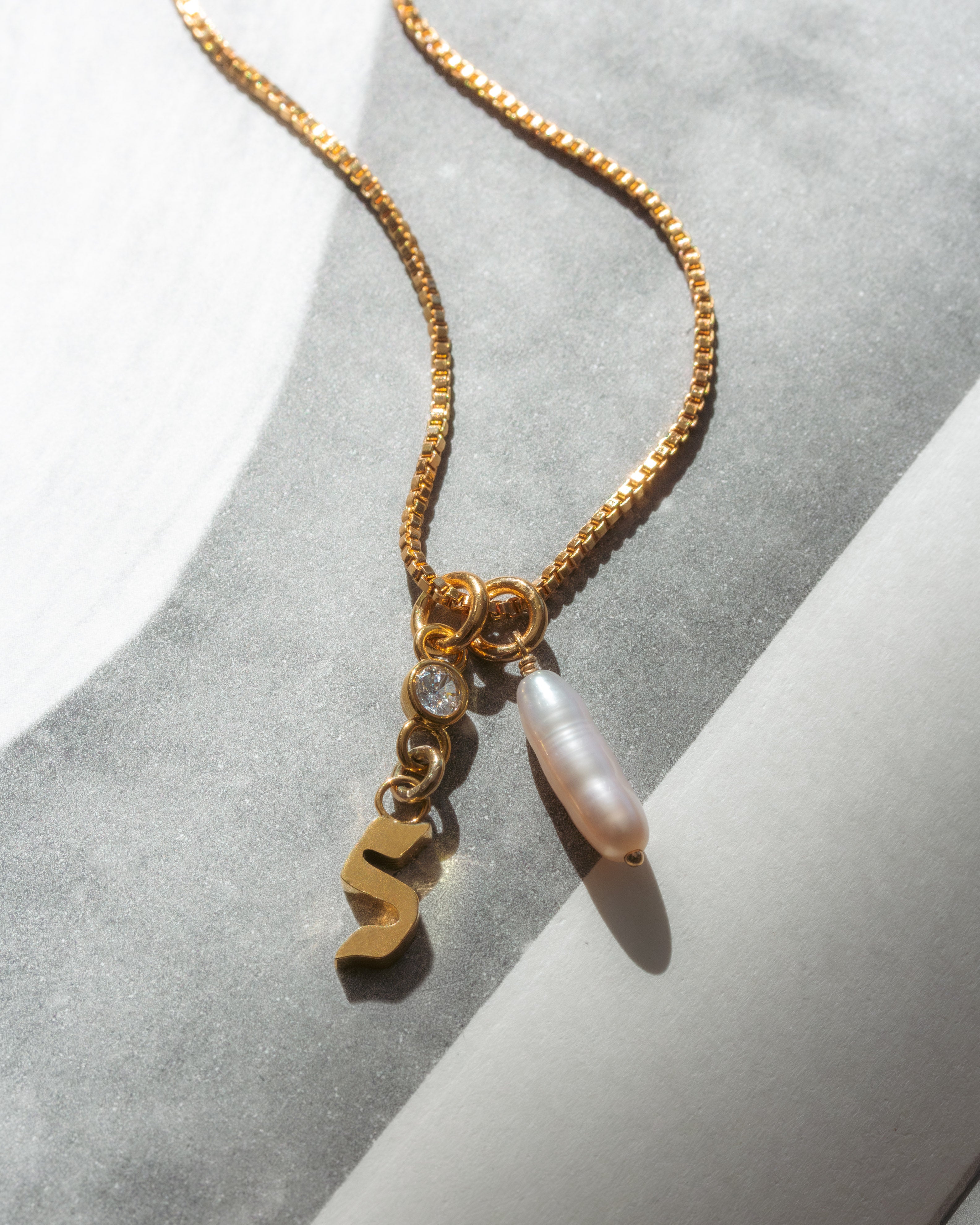 Gold box chain necklace with an S initial charm, a round clear crystal, and an long pearl pendant.