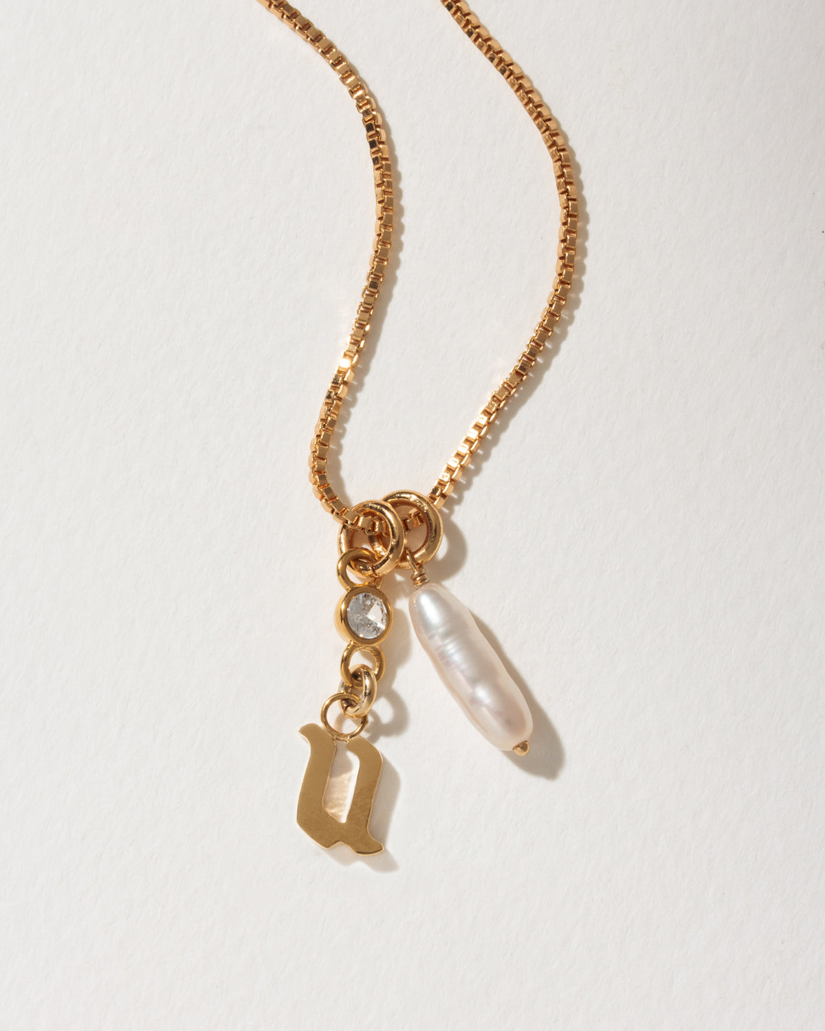Gold box chain necklace with a gothic initial U charm, a small clear crystal, and a baroque pearl.