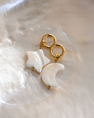 Two earring charms, one in the shape of a star and the other a crescent moon, both with a mother of pearl design, attached to gold-colored hoop earrings.