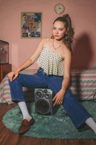 A young woman sits on a speaker upon a green rug in a pink bedroom, wearing a gemstone charm necklace, a cream polka-dot spaghetti strap top, blue jeans, white socks, and brown loafers. 