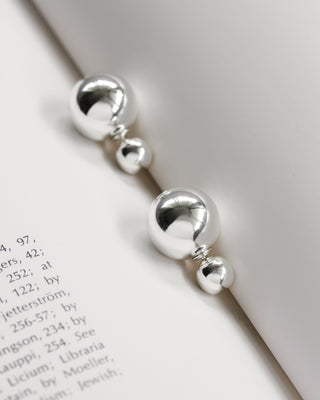 A pair of silver earrings with a polished finish, featuring a front post design with an orb backing, displayed on a white surface with text in the background.