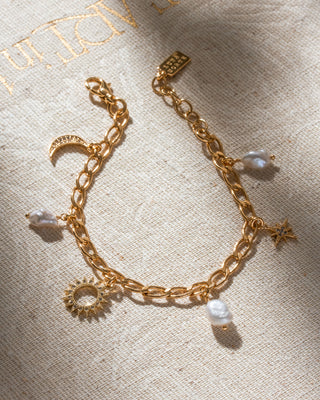 Gold plated bracelet with cz elements and sun, moon, star charms, and pearl beads on a textured beige background