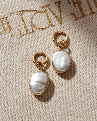 Two oval-shaped pearl charms with gold-colored hoop closures, placed on a beige fabric surface.