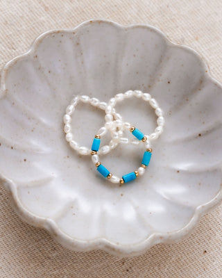 Three stretch rings featuring cultured freshwater pearls and genuine turquoise, displayed stacked on a scalloped white dish.