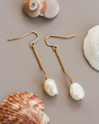 Pearl earrings with gold chains on a surface with seashells.