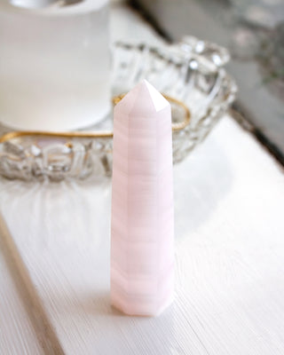 Pinkish striped calcite gemstone obelisk tower on a white window sill with out of focus object in the background.