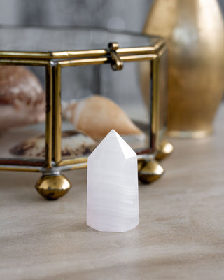 A white calcite gemstone tower placed on a flat surface on a table with other curios, displaying its smooth, faceted shape.