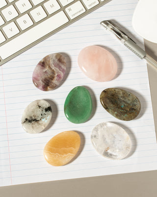 Six polished tumbled stones in various colors and patterns are arranged on lined notebook paper next to a keyboard and pen.