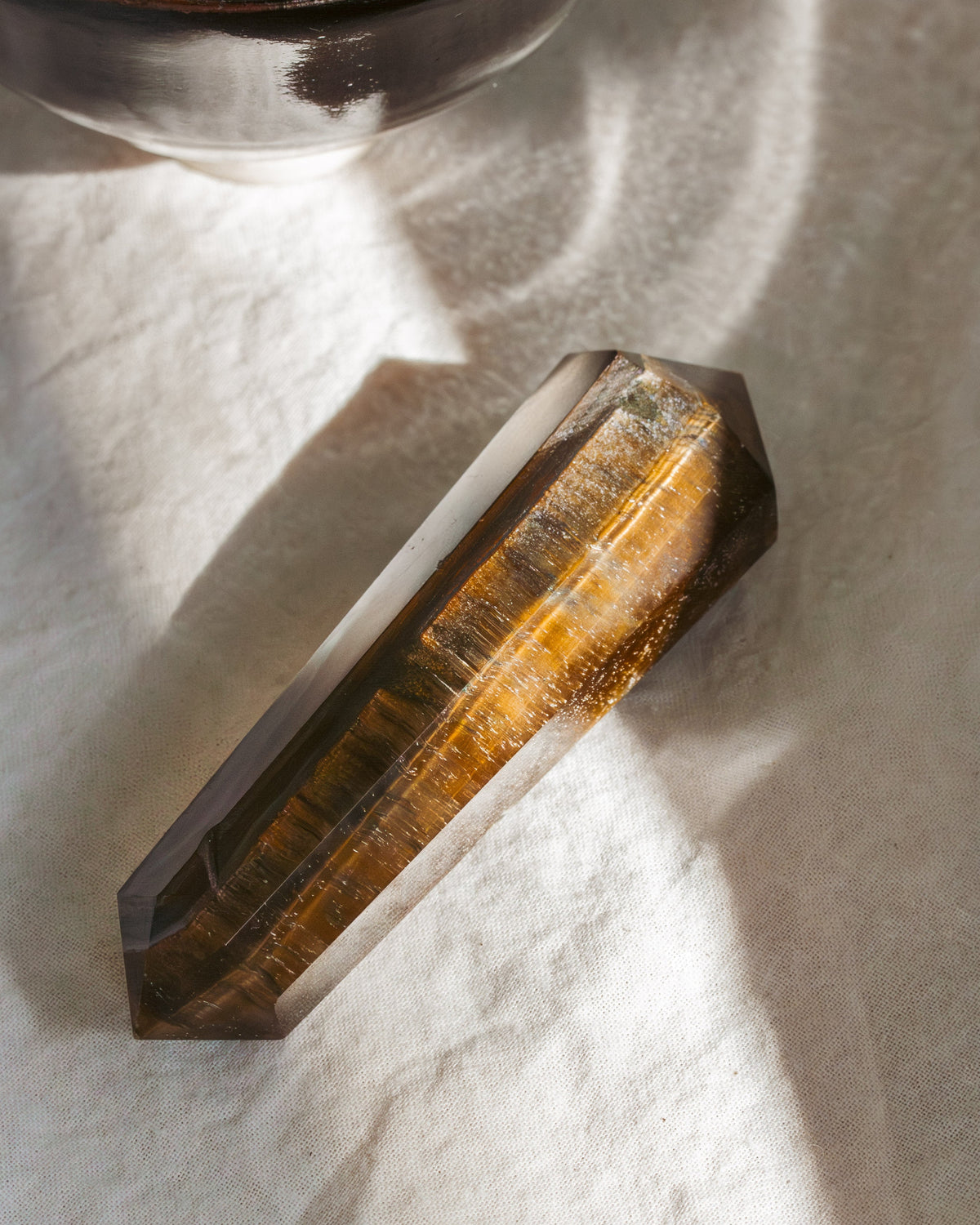 A faceted tiger's eye crystal point resting on a textured white fabric surface, with a dark ceramic bowl partially visible in the top left corner under warm, directional sunlight.