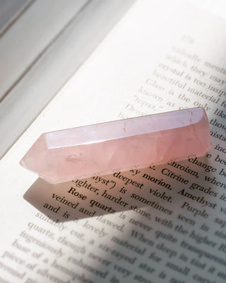 A polished pink rose quartz crystal point resting on an open book with printed text, casting a soft shadow.