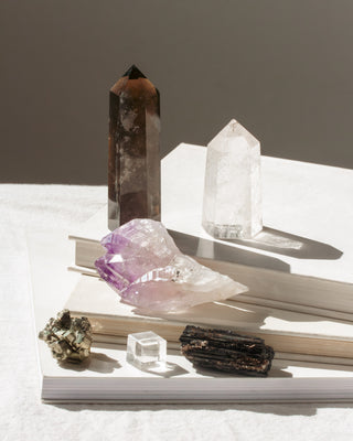 Variety of gemstone wands and rough gems stacked on top of books.  From the metallic shimmer of pyrite and the rich depth of smoky quartz to the grounding texture of black tourmaline and the crystalline formations of amethyst.