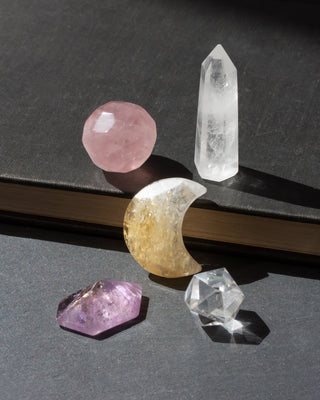 Gemstone wand, moon, and other geometric shapes resting on a table. The Good Energy Gemstone Curio Set features 5 genuine gemstone curios, each selected for its unique form and visual appeal.

This playful assortment includes a faceted geometric rose quartz sphere, mini clear quartz tower, citrine crescent moon palm stone, geometric amethyst specimen, and a octahedron clear quartz crystal. 