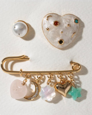 A gold-tone pin set, featuring a heart-shaped enamel brooch with small colored gemstones, a faux pearl stud, and a safety-pin style charm bar with dangling heart, crescent moon, star, and flower charms.