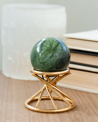A green serpentine gemstone sphere on a gold-colored geometric stand, placed on a wooden surface with books in the background.
