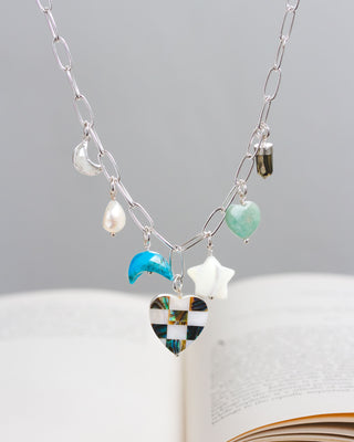 A silver chain necklace with a variety of charms including a mother-of-pearl heart, abalone, and other gemstones, displayed against a book backdrop.