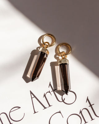 Two smoky quartz crystal charms with gold-colored caps and loops, lying on a surface with a shadow cast over them.