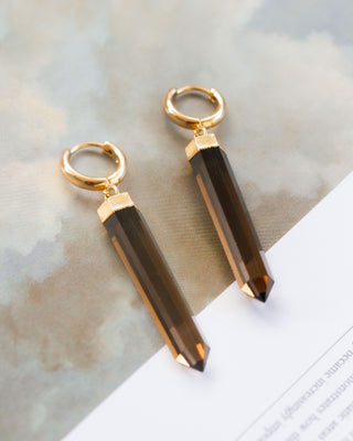Two smoky quartz huggie hoop earrings with gold-colored caps and hooks, displayed against a neutral background.