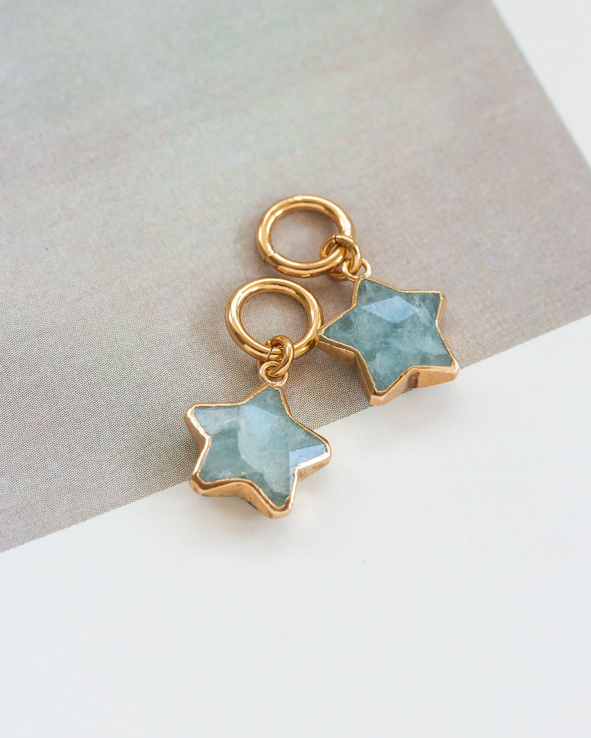 Two earring charms designed with aquamarine star shapes, attached to gold hoop closures