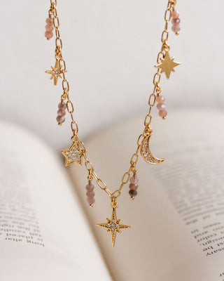 A gold-toned necklace with celestial charms including a crescent moon and stars, and coffee moonstone beaded drops, displayed against a backdrop of an open book.