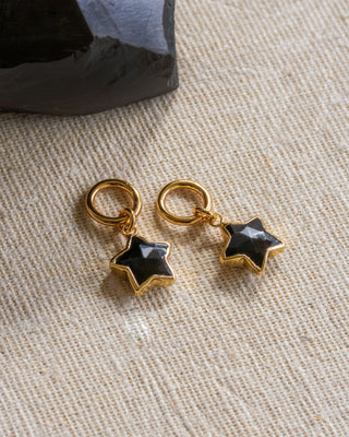 Two hoop earrings with star-shaped black onyx gemstones attached to them, placed on a textured fabric.