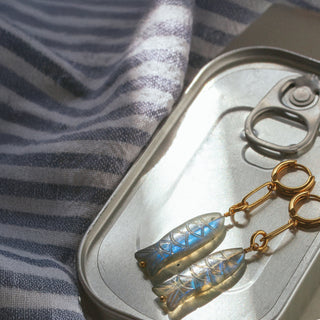 Gold fish-shaped earrings on a metal can with striped fabric background