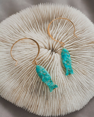 Gold hoop earrings with turquoise fish charms on a coral surface