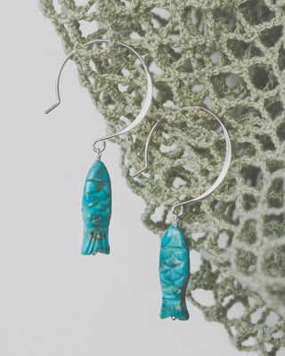 Silver Turquoise fish-shaped earrings on a textured green fabric background