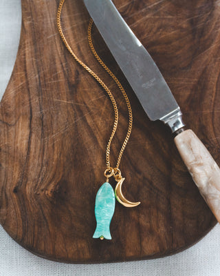 Gold necklace with a turquoise fish pendant and gold crescent moon charm on a wooden surface with a knife.