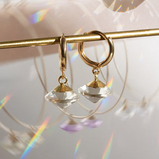 Gold Mini huggie hoop with quartz charm hanging on gold rod infront of rainbow refractions and blurred background image