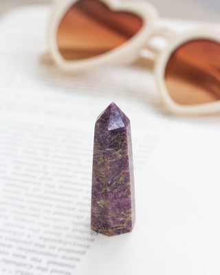 Lepidolite mini tower obelisk standing on a book page with heart shaped sunglasses in the background.