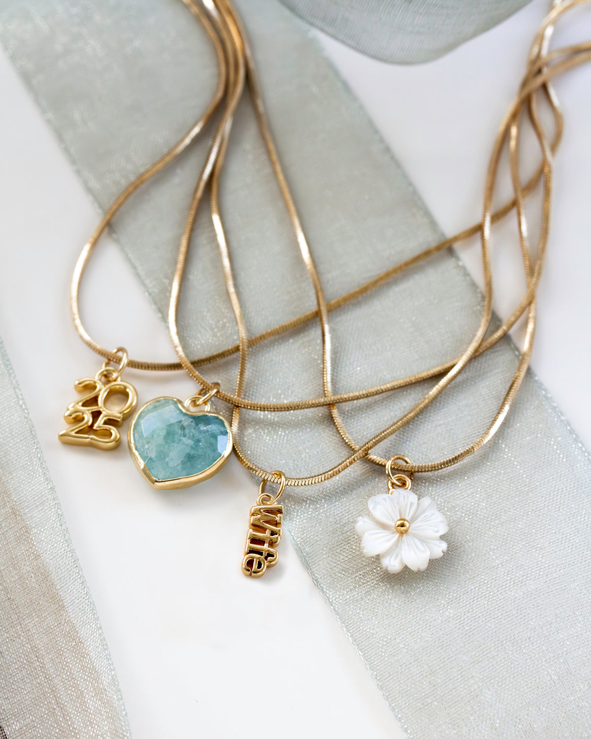 Gold necklaces with various pendants, including aquamarine heart, 2025, Wife, and mother of pearl floral charms, on a light fabric background.