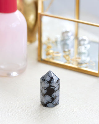 A mini snowflake obsidian gemstone tower with a black background and gray dusting, placed on a flat surface.