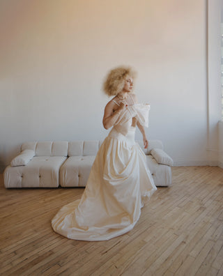 Woman in a flowing bridal gown stairs out of a window. She is standing on a hardwood floor with a white couch in the background.