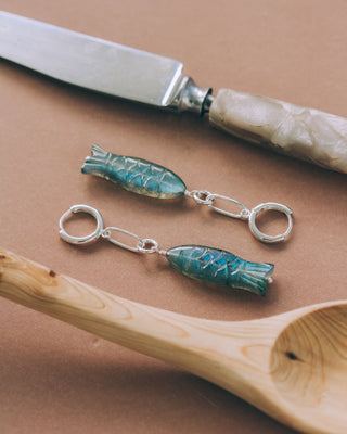 Two fish-shaped charm earrings next to a knife and wooden spoon on a brown background
