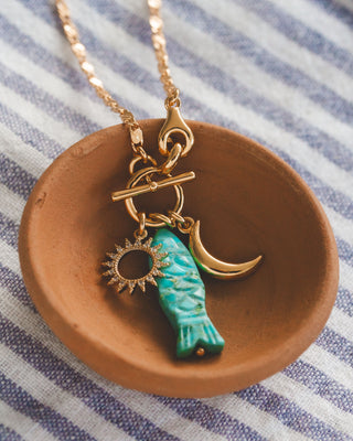 Gold necklace with turquoise fish charm, sun charm, and moon charm on a brown dish with striped fabric background