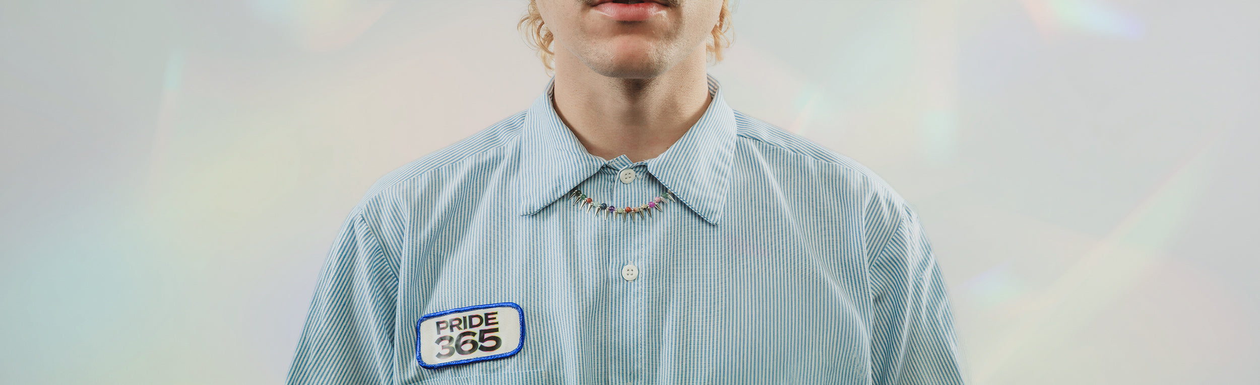 Person wearing multicolor beaded and silver spiked necklace and a light blue striped shirt with a patch showing Pride 365 collection logo, on a light rainbow background