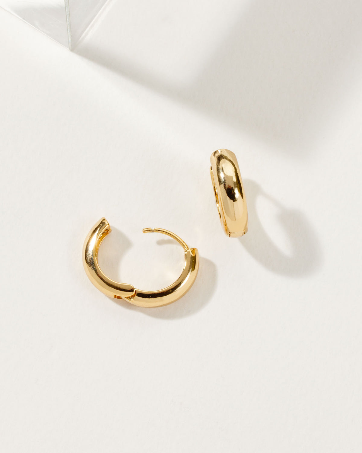 Two shiny gold Huggie Hoop Earrings on a white surface.