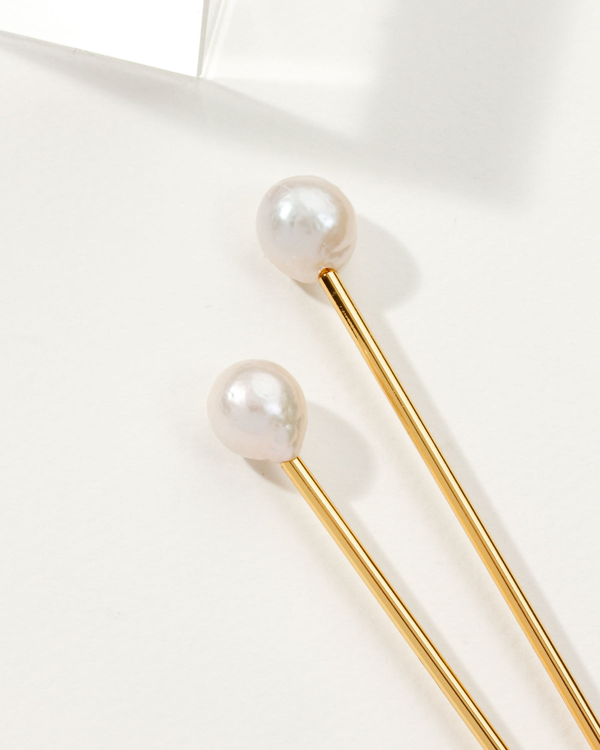 Two pearl hair pins with gold shafts lay diagonally on a white surface.